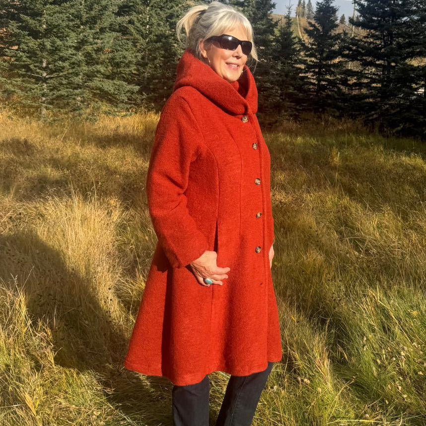 A woman wearing a bright red wool coat, black pants, and sunglasses stands in a grassy field with pine trees and autumn foliage in the background.